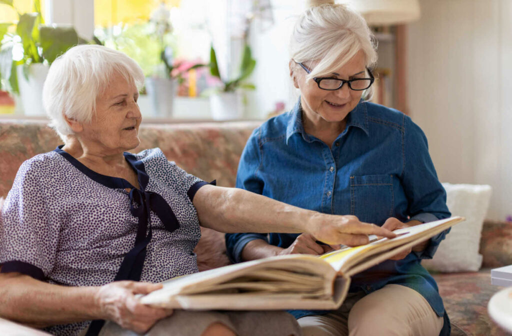 Two senior female is sitting beside each other on a flower-designed couch while they are looking into a photo album.