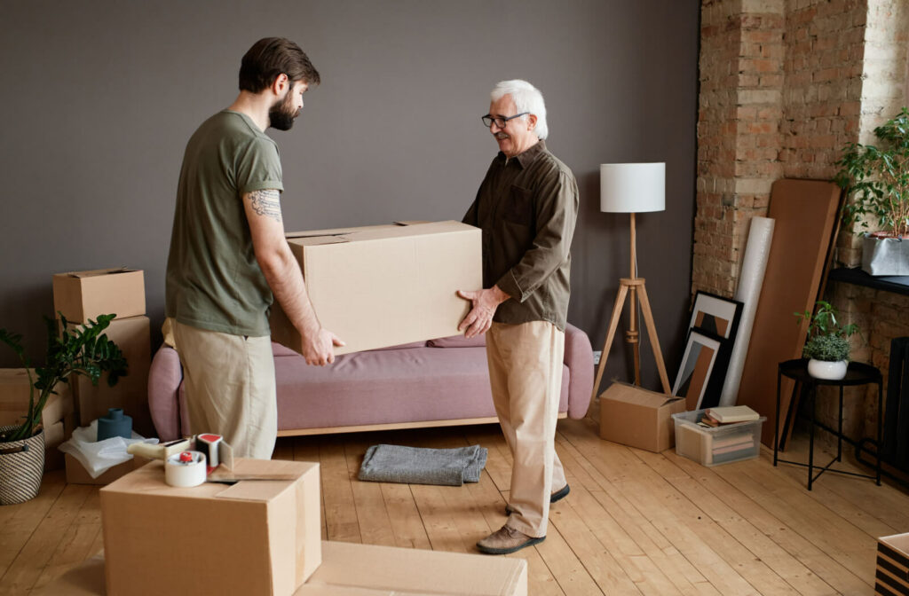 A young man helping a senior man move cardboard boxes in a living room.