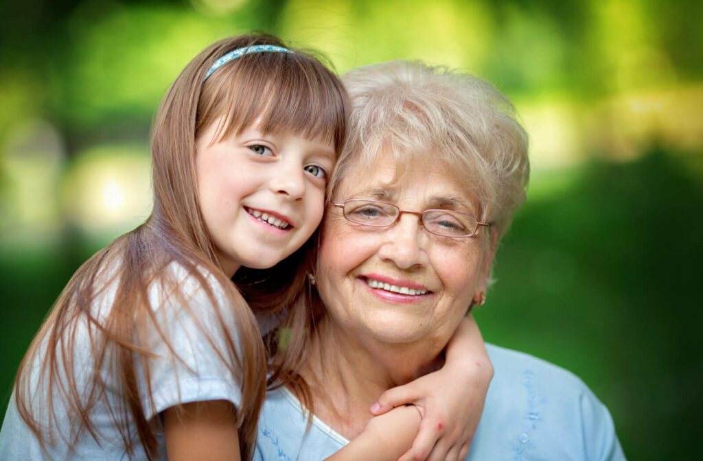 An older adult woman with her granddaughter in a park, hugging her, smiling and looking directly at the camera